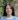 Head and shoulders of a woman with long dark hair parted in the centre, looking directly at the camera. She is wearing a light blue jumper with a white T shirt just seen underneath. Behind her is a glimpse of old red brick wall with ivy growing on it.